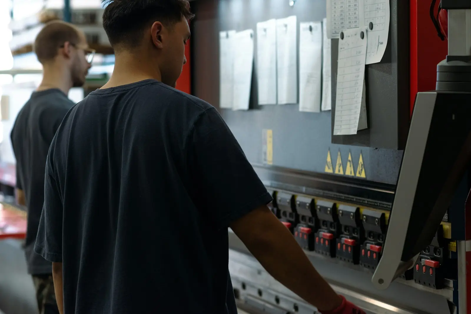 Two operators at a press brake, the work sheets pinned above the machine.